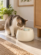 Chat buvant dans la fontaine en céramique blanche Cérélia installée près d’un meuble en bois.